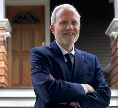 David Ruzumna in a navy suit with arms crossed, standing outside on a home porch