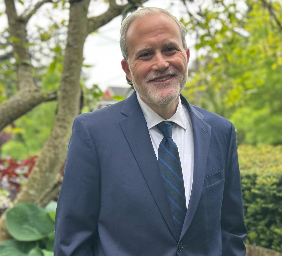 David Ruzumna smiling in a navy suit and striped tie, photographed with a garden backdrop